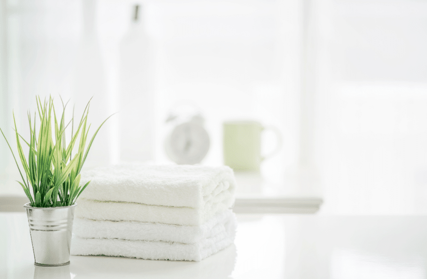 White towels on white counter with green plant in silver container, in background and blurred is white coffee cup and white alarm clock