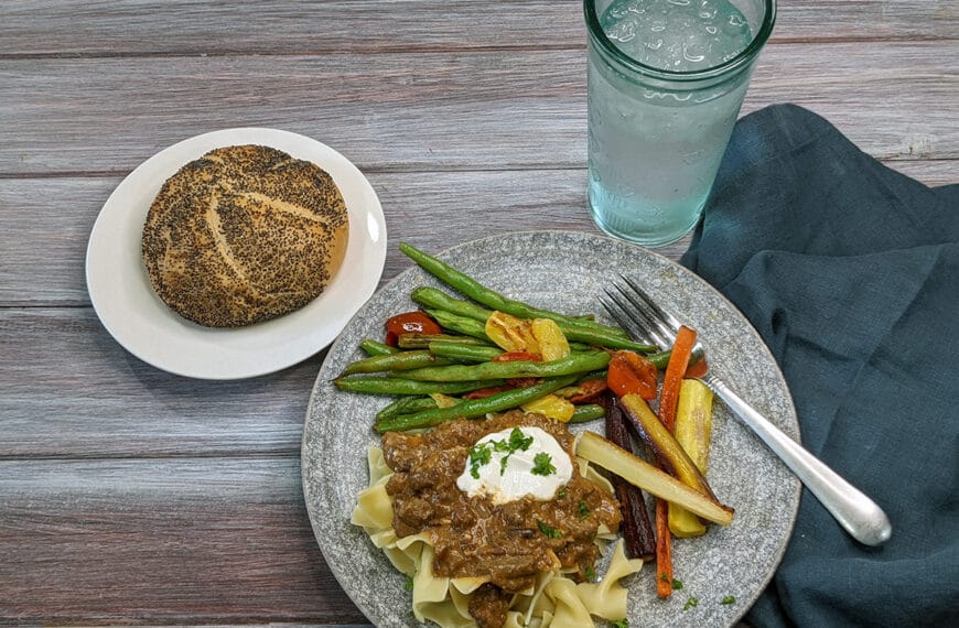 A roll with poppy seeds on a white plate, a plate of ground beef stroganoff over egg noodles with a dollop of sour cream, roasted carrots, and pan seared green beans with multi-colored grape tomatoes, a water glass and a blue napkin